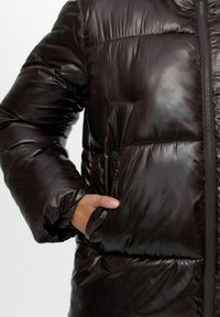 Tumepruun puffer-jakk, millel on särav viimistlus, varustatud lukuga ja esikoti näidikuga. Kude on sile, kergelt peegeldava tekstuuriga.