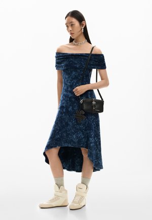 Mujer con vestido azul de terciopelo con escote off-shoulder y dobladillo asimétrico, zapatillas beige altas, calcetines grises y bolso cruzado negro con detalle de flores.