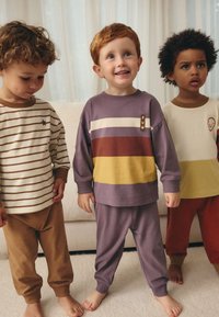 Trois enfants portent des vêtements d'intérieur coordonnés : un t-shirt rayé avec une étoile, un t-shirt violet avec des rayures multicolores, et un t-shirt crème avec un visage souriant.