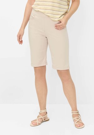 Vrouw draagt beige knielange shorts, gestreepte geel-witte top en beige platte sandalen met bandjes, staand met één hand op de heup.