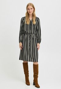 Schwarz-weiß gestreiftes Kleid mit einem Bindeband in der Taille, langen Ärmeln und einem entspannten Schnitt, ergänzt durch braune kniehohe Stiefel.