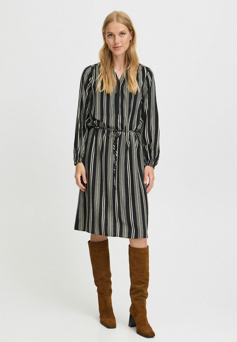 Schwarz-weiß gestreiftes Kleid mit einem Bindeband in der Taille, langen Ärmeln und einem entspannten Schnitt, ergänzt durch braune kniehohe Stiefel.