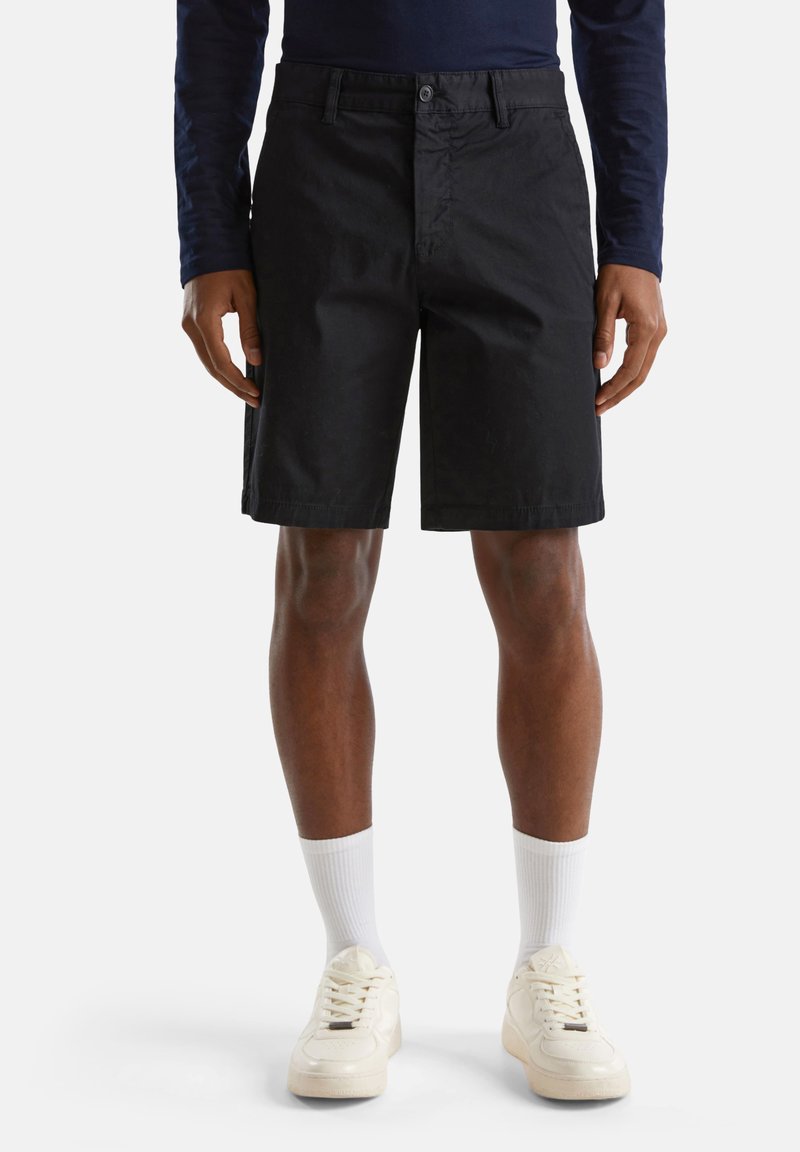Shorts noirs en tissu, avec une coupe classique, fermeture à bouton et poches latérales. Portés avec des chaussettes blanches et des baskets crème.