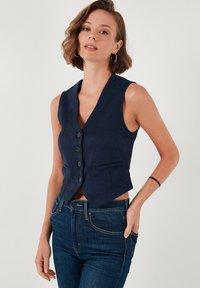 Navy plavi suede prsluk s V-izrezom, zatvaranjem na pet dugmadi, dva bočna džepa i skraćenim dizajnom. Karakterizira ga gladak tekstur i krojen fit.