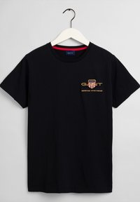 T-shirt noir à manches courtes avec un col rond, accroché à un cintre blanc, avec un petit logo orange et beige sur la poitrine.