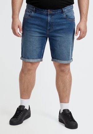 Un homme porte un short en jean bleu à revers, un tee-shirt noir, des chaussettes blanches et des baskets noires sur un fond uni.