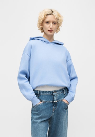 Άτομο με κοντά σγουρά ξανθά μαλλιά που φοράει ανοιχτό μπλε φούτερ με κουκούλα και ψηλόμεσα μπλε τζιν, με τα χέρια στις τσέπες, στέκεται μπροστά σε απλό φόντο.