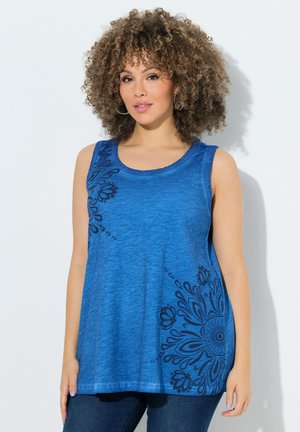 Kobieta o kręconych włosach, ubrana w niebieski top bez rękawów z czarnymi kwiatowymi wzorami, stojąca na białym tle.