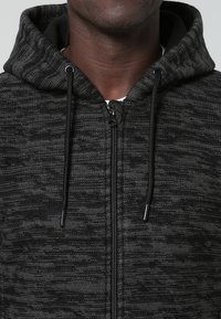 Närbild av en person som bär en mörkgrå strukturerad zip-up hoodie med svarta snörningar och huvan uppe, vilket visar halsen och nedre delen av ansiktet.