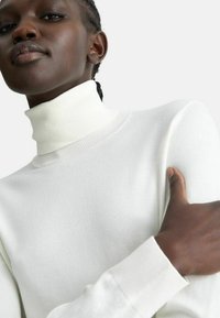 Maglione bianco a collo alto con colletto e polsini a coste, realizzato in un tessuto a maglia liscia. Inquadratura ravvicinata che mostra la trama e la vestibilità.