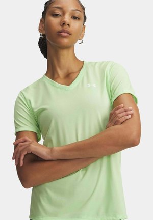 Giovane donna con capelli intrecciati che indossa una maglietta verde chiaro a maniche corte con scollo a V, braccia incrociate, espressione neutra, sfondo chiaro uniforme.