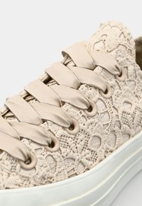 Beżowe sneakersy z koronką z teksturowanym, skomplikowanym wzorem, płaskimi sznurowadłami i białą gumową podeszwą z subtelnymi żółtymi akcentami.