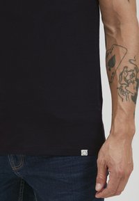 Black cotton t-shirt with a crew neckline. Features a small logo tag at the bottom hem. Worn with dark blue jeans. Smooth texture.