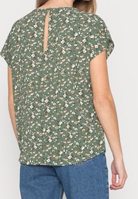 Zielona bluzka z krótkim rękawem w białe i pomarańczowe kwiatowe wzory, z dekoltem w kształcie łezki na plecach i luźnym, swobodnym krojem.
