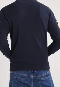 Sudadera azul marino hecha de tejido suave, con cuello redondo, mangas largas y puños acanalados; parte trasera lisa sin logotipos ni estampados visibles.