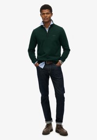 Grøn strikket pullover med halvlynlås krave over en lys blå skjorte, parret med mørke jeans og brune sko.