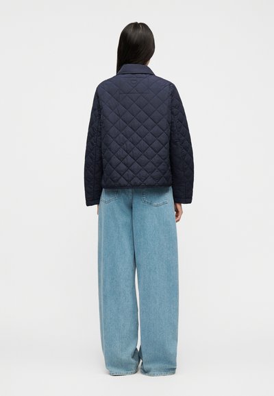 Άτομο με μακριά σκούρα μαλλιά που φοράει σκούρο μπλε καπιτονέ μπουφάν και χαλαρό ανοιχτό μπλε τζιν, στέκεται με την πλάτη γυρισμένη σε απλό φόντο.