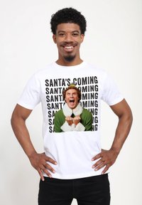 Biała bawełniana koszulka z kolorową grafiką postaci w zielonym stroju z białymi akcentami, tekst brzmi "ŚWIĘTY MIKUŁAJ NADCHODZI."