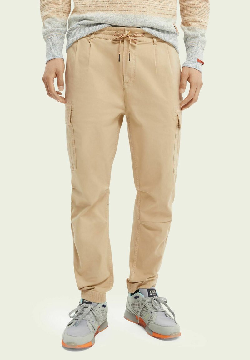 Mand iført beige cargo-bukser med snor i taljen og lommer, grå og orange sneakers samt en lysefarvet sweater.
