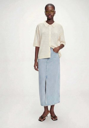 Kvinde iført gultonede briller, cremefarvet kortærmet strikket cardigan, lyseblåt højtaljet midi denimnederdel med frontslids og sorte sandaler med hæl.