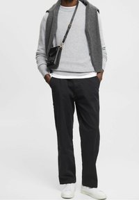 Grau gestrickter Pullover unter einer dunkelgrauen ärmellosen Strickjacke, schwarze Hose und weiße Sneakers. Kleine schwarze Umhängetasche.
