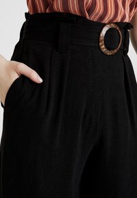 Black trousers with a high waist, pleats, side pockets, and a decorative round brown belt buckle. Smooth fabric texture.