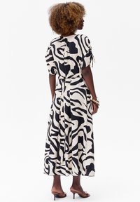 Maxikleid mit Schwarz-Weiß-Zebramuster, tailliert, mit kurzen Ärmeln und weichem, fließendem Stoff; getragen mit braunen Sandalen mit Absatz.