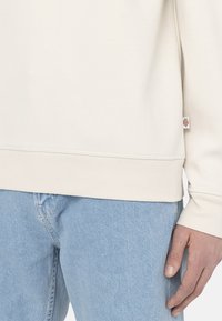 Felpa color crema con bordo e polsi a costine, realizzata in tessuto morbido. Un piccolo logo è visibile vicino al bordo inferiore. Indossata con jeans azzurri.