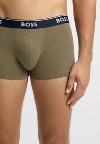 Man wearing olive green boxer briefs with a navy waistband featuring white "BOSS" branding, standing with his right hand relaxed by his side.