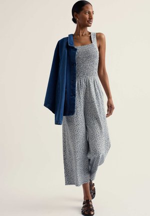 Femeie purtând un salopetă fără mâneci cu imprimeu floral albastru și sandale închise la culoare, cu o jachetă din denim albastru așezată pe un umăr.