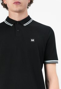 Black polo shirt featuring white-striped collar and cuffs, three-button placket, textured fabric, and a small branded patch on the chest.