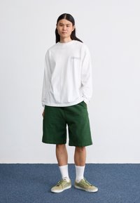 Chemise blanche à manches longues avec un petit logo noir, associée à un short vert, des chaussettes blanches et des baskets beiges. Tenue simple et décontractée.