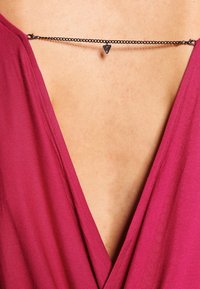 Schwarze Metalldraht-Halskette mit einem kleinen dreieckigen Anhänger, getragen über einem tiefen V-Ausschnitt aus festem maroonfarbenem Stoff.