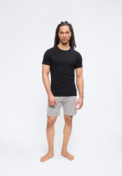 Άνδρας με μακριά dreadlocks που στέκεται ξυπόλυτος, φορώντας μια εφαρμοστή μαύρη μπλούζα και γκρι σορτς, μπροστά σε ένα απλό φωτεινό φόντο.