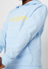Ljusblå hoodie med en stor gul "LACOSTE"-logotyp, med ärmar i rutmönster och en framficka. Tillverkad av mjukt tyg.
