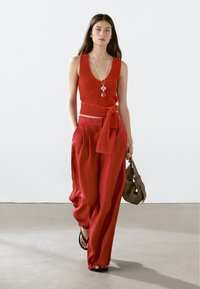 Röd stickad kort topp med djup V-ringning, kombinerad med lösa röda byxor med knytliv, kompletterad med en brun handväska och sandaler.