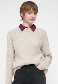 Suéter beige de canalé con escote redondo, combinado con una camisa negra de cuello burdeos por debajo, usado con pantalones negros.