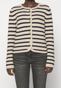 Raidoitu neuletakki vaaleassa kerman ja laivastonsinisen sävyssä, jossa on pyöreä kaula-aukko, kultaiset napit, ribattu pinta ja istuva malli.