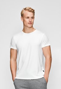 Hvid kortærmet t-shirt med rund hals, blødt stof og slim fit, parret med grå bukser. Har et lille sort logo nær sømmen.