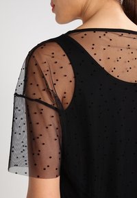 Primer plano de una mujer que lleva una blusa negra transparente con pequeños lunares negros sobre una camiseta negra sólida, mostrando el hombro y la parte superior de la espalda.