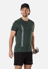 Homme portant un T-shirt vert foncé "Swedemount", un short noir et une casquette, tenant une bouteille d'eau, regardant sur le côté, debout devant un fond uni.