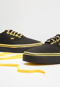 Mustat kangaskengät, joissa on keltaiset nauhat ja reunus, sekä tasainen kumipohja. Nauhoissa on "VANS"-brändin ompelu.