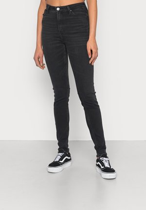 Persoon draagt zwarte skinny jeans en zwart-witte sneakers, staand tegen een effen witte achtergrond.