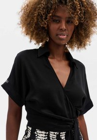 Zwarte wikkelblouse met een V-hals en opgerolde mouwen, gemaakt van een gladde stof. Voorkant gestrikt op de taille, gecombineerd met een gestreepte rok.