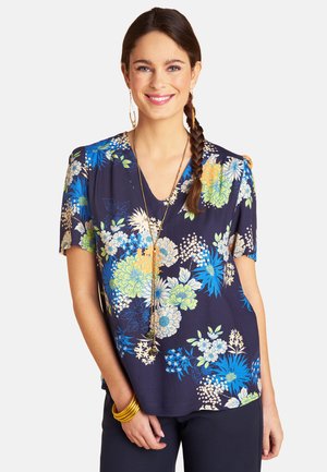 Woman with braided hair smiling, wearing a navy floral short-sleeve top, long gold necklace, gold hoop earrings, and gold bracelets.