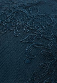 Donkerblauwe stof met complexe bloemembroideries met verhoogde, textuurelementen tegen een lichte, sheer achtergrond.