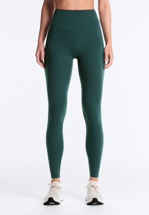 Frau trägt dunkelgrüne Leggings mit hoher Taille und weiße Turnschuhe, steht mit entspannten Armen seitlich am Körper vor einem einfarbigen Hintergrund.