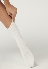 Witte ribbenkatoenen sok, midi-kuitlengte, met een gladde textuur en elastische bovenkant, die door een hand omhoog wordt getrokken.
