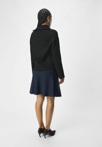 Suéter negro de canalé con cuello y ajuste entallado, combinado con una falda acampanada azul marino. Unos zapatos de tacón negro de punta afilada completan el conjunto.