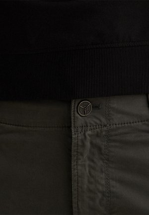 Primer plano de pantalones verde oscuro con cierre de botón metálico con marca debajo de una pretina negra acanalada.